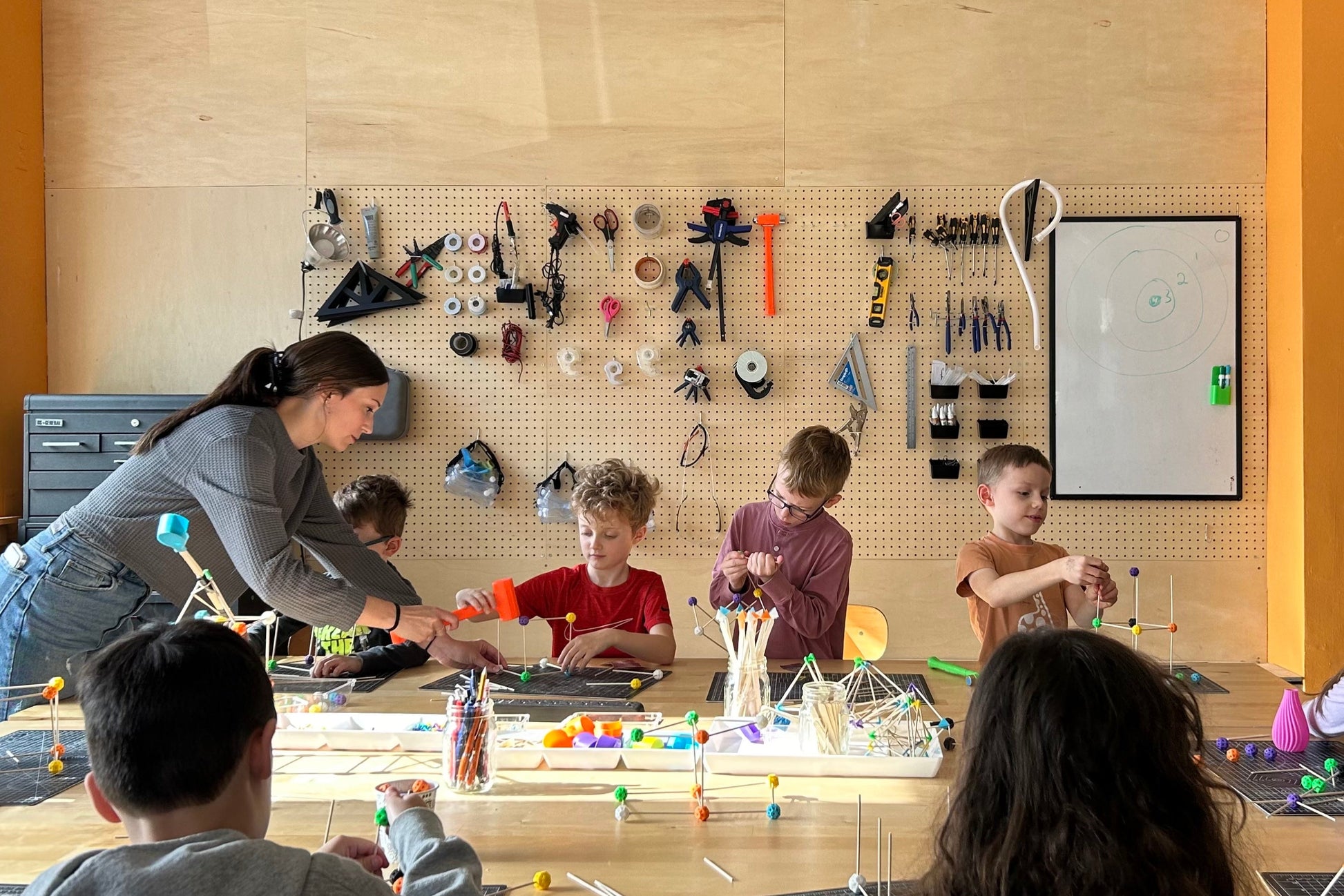 Children and a teacher engaged in an educational activity at a table with various tools and materials on a pegboard wall.